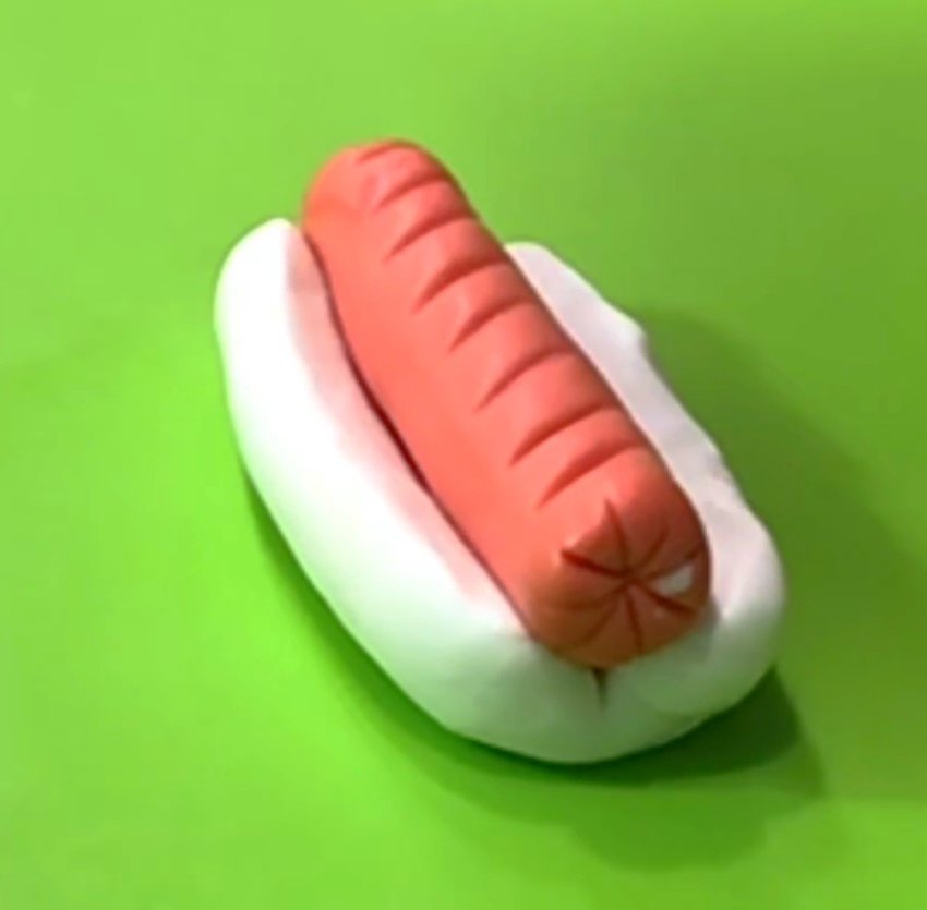 Roll out white fondant and shape like a hot dog bun. Slice it lengthwise with an X-Acto knife (but not all the way through), then fold it open. Mix fondant colors to make your choice of color for the hot dog. Mold to create the shape, and texture with an X-Acto knife and a clay tool.